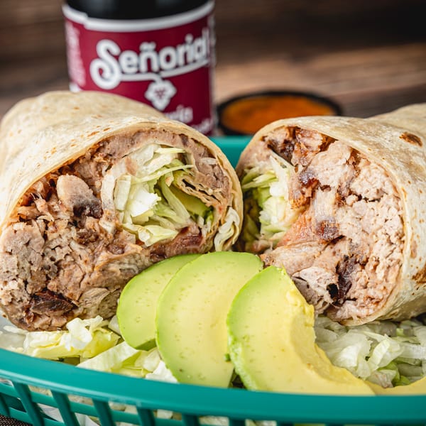 Two halves of a large burrito with meat, lettuce, and avocado slices in a green basket.