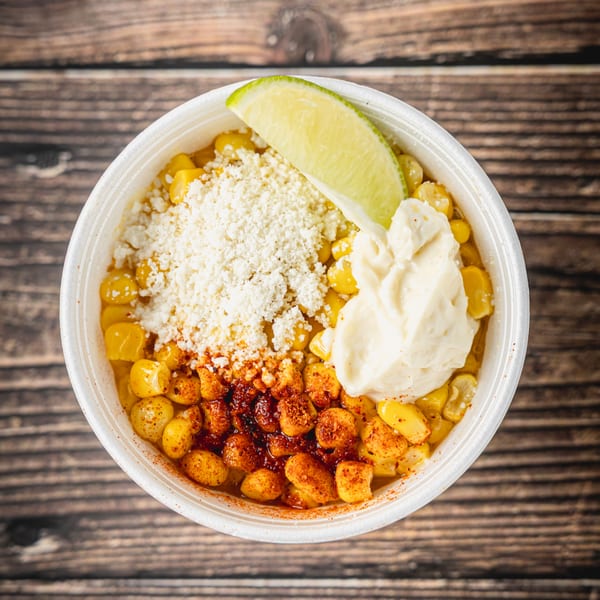 Elote in a white cup, topped with cheese, mayonnaise, chili powder, and a lime wedge.