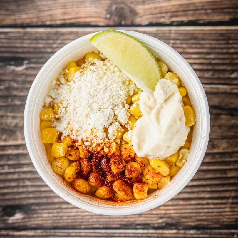 Elote in a white cup, topped with cheese, mayonnaise, chili powder, and a lime wedge.