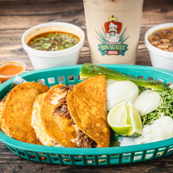 Taquitos Don Agaves birria tacos with consome, beans, and a branded drink on a wooden table.