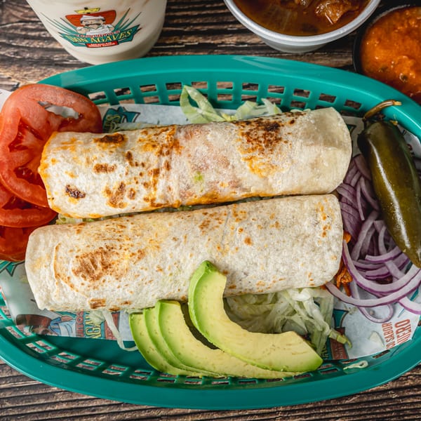 Two golden-brown burritos in a green basket with avocado, tomato, red onion, and a jalapeño.