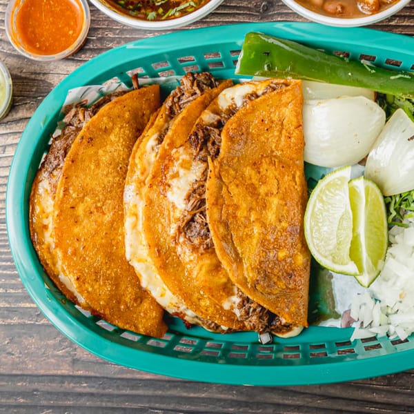 A teal basket filled with four crispy birria tacos, lime wedges, grilled peppers, and onions.