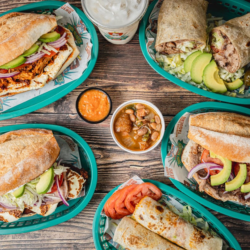 Overhead view of Taquitos Don Agaves burritos, tortas, beans, and salsa on a wooden table.
