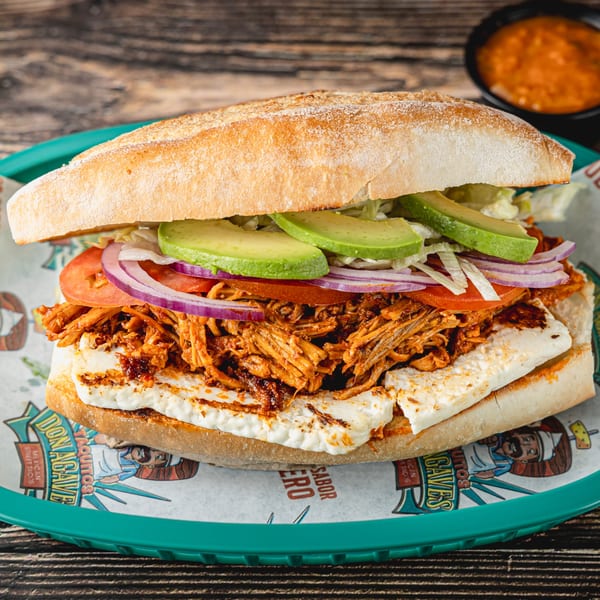 A large torta sandwich with shredded meat, cheese, avocado, tomato, onion, and lettuce.