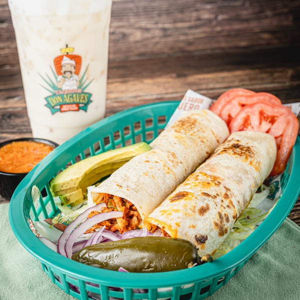 Two burritos in a green basket with avocado, red onion, tomato, jalapeño, and salsa, with a Don Agaves drink.